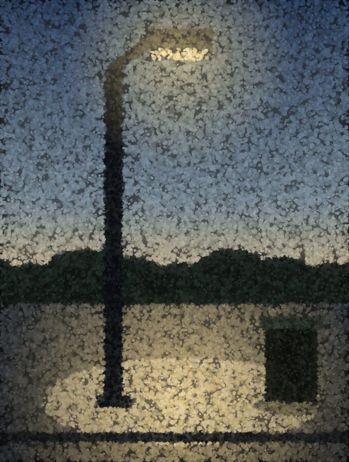 A single street lamp at dusk casting a bright pool of light on a dark pavement, with a bin at the edge of it.