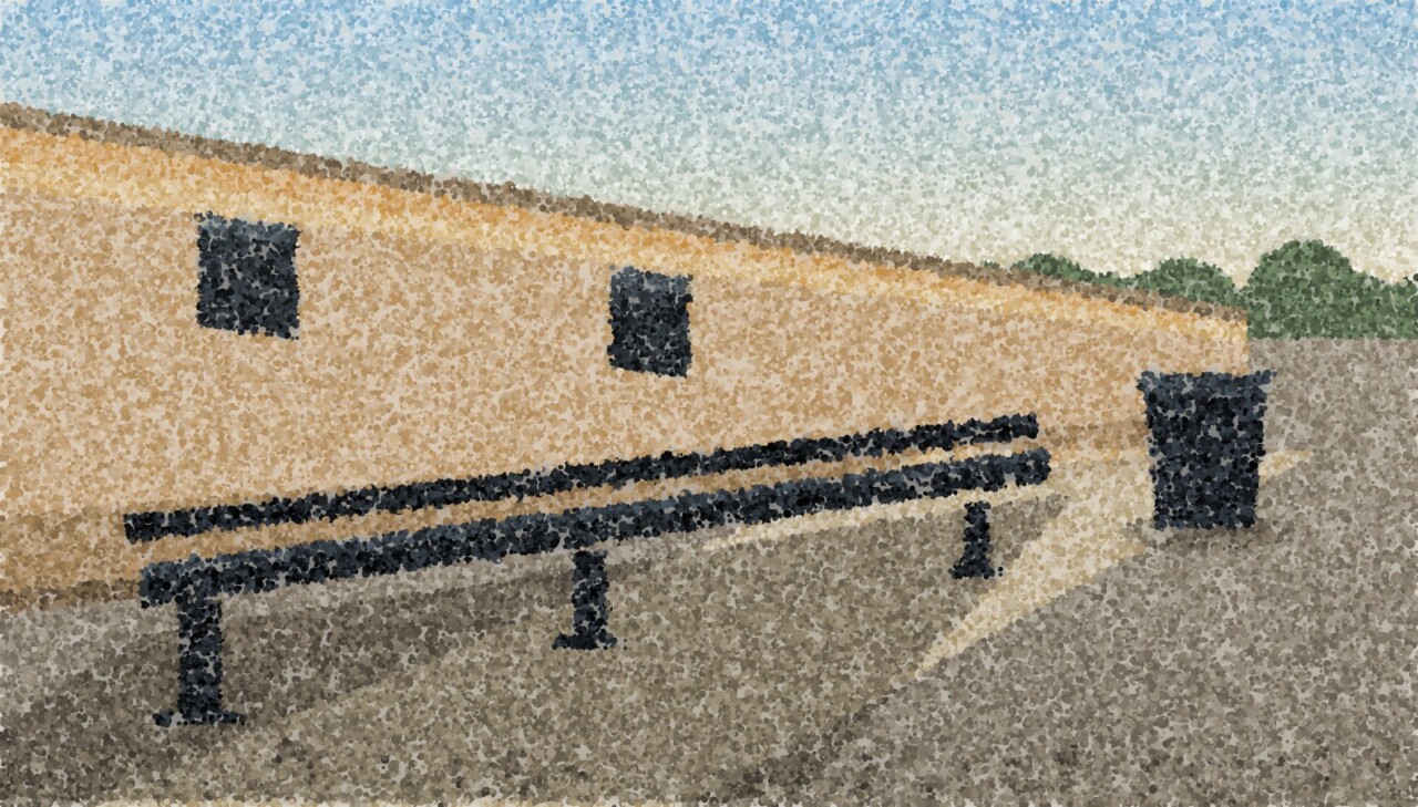 A long bench standing against a high yard wall in low sun, a bin at the end of it, the tarmac empty.