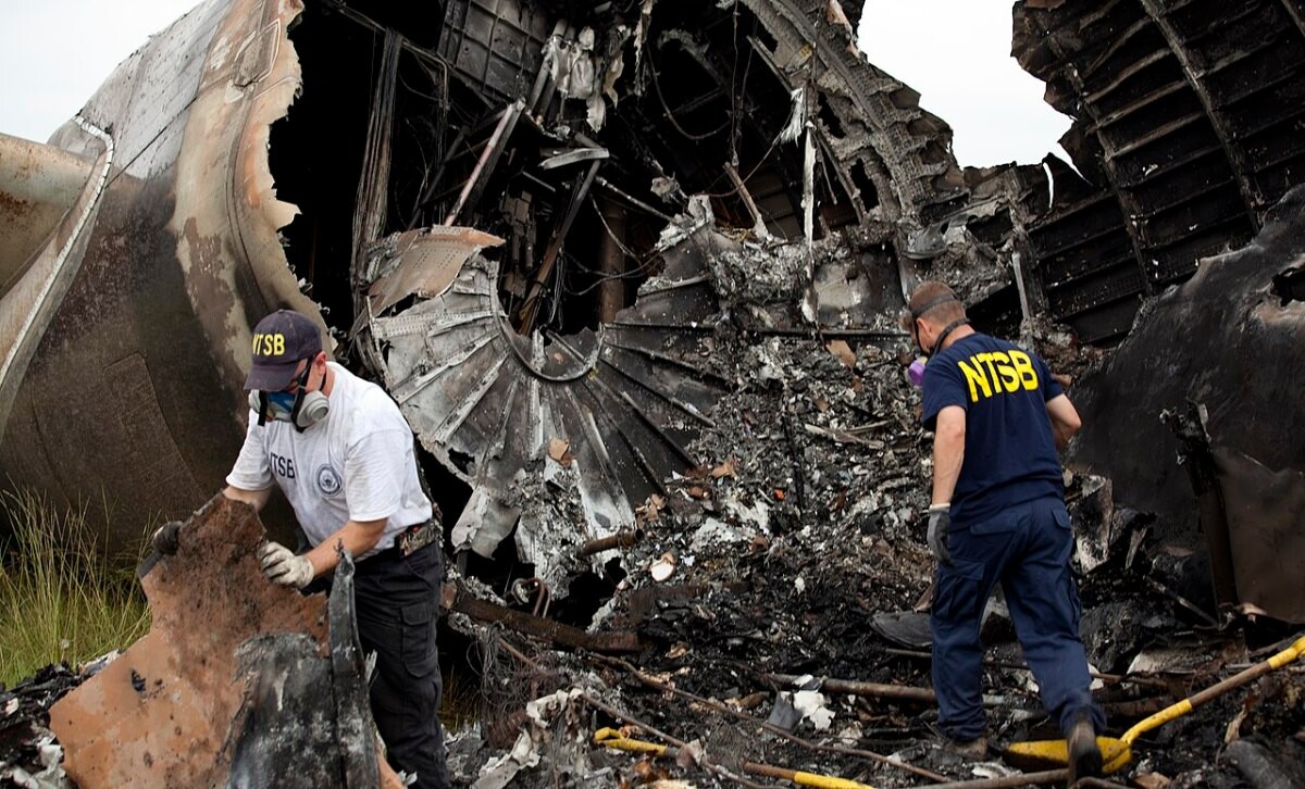 Two NTSB investigators examining twisted aircraft wreckage in a field
